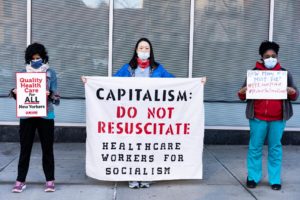 Healthcare workers in masks hold up three protest signs and banners: Quality Health Care for All New Yorkers, Captialism: Do Not Resucitate: Healthcare Workers for Socialism, How Many of us must die? #PPEoverprofit #ProtecttheFrontLine