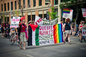 Two people carry a banner in a Pride march which says "Queers Against Israeli Apartheid" and which has on one side the flag of Palestine and on the othernd the rainbow flag. Behind them another banner reads "Free Palestine"