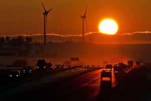 A sun through dark haze with a highway and cars with two windmills in the foreground