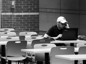 A man working alone on a laptop at a white table in a room with rows of identical white tables.