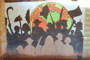 Silhouettes of demonstrators with flags and agricultural equipment in front of a representation of the globe bordered with the phrase Movimento Sem Terra painted on a wall
