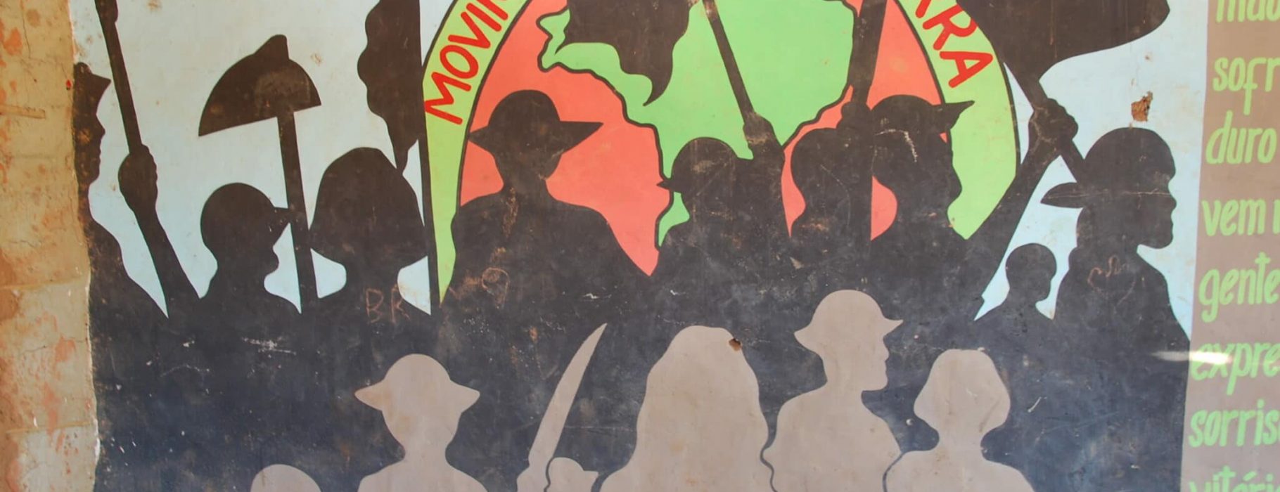 Silhouettes of demonstrators with flags and agricultural equipment in front of a representation of the globe bordered with the phrase Movimento Sem Terra painted on a wall