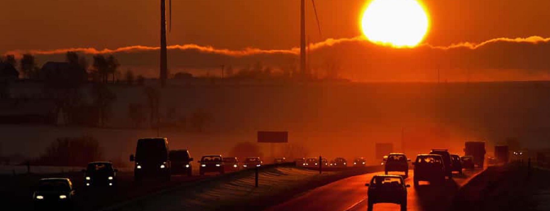 A sun through dark haze with a highway and cars with two windmills in the foreground