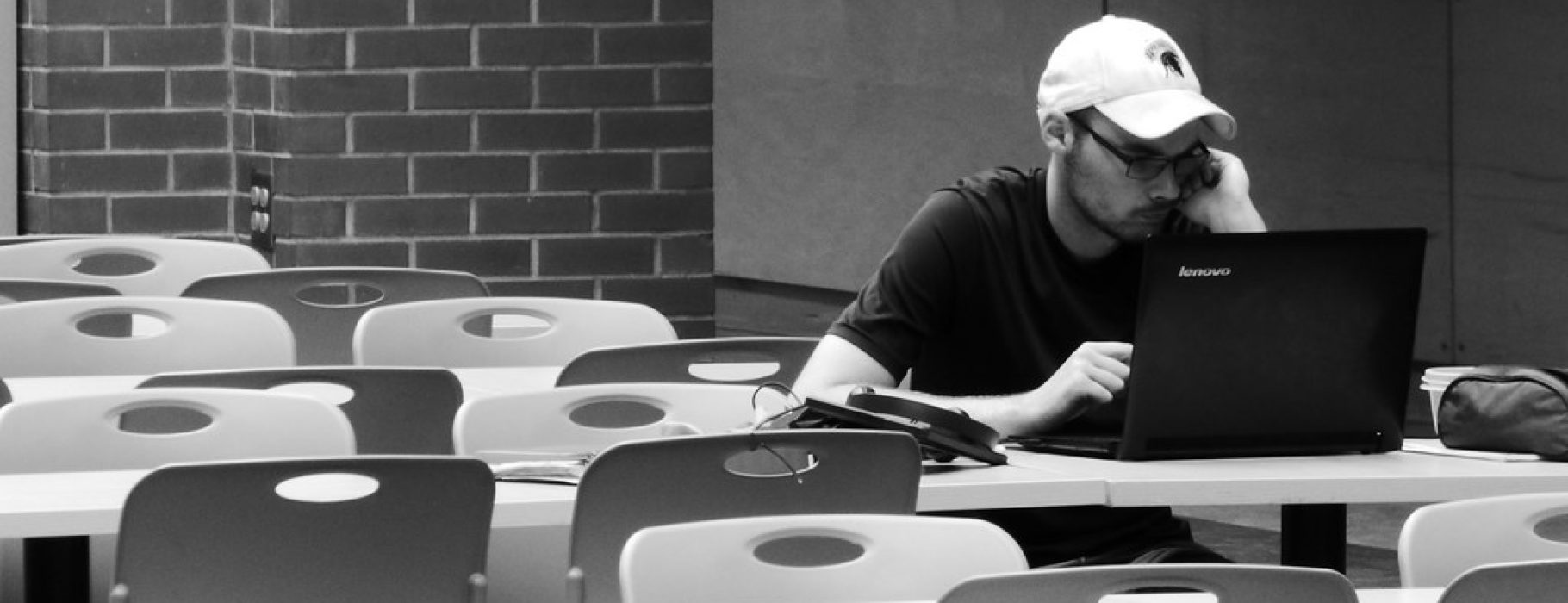 A man working alone on a laptop at a white table in a room with rows of identical white tables.