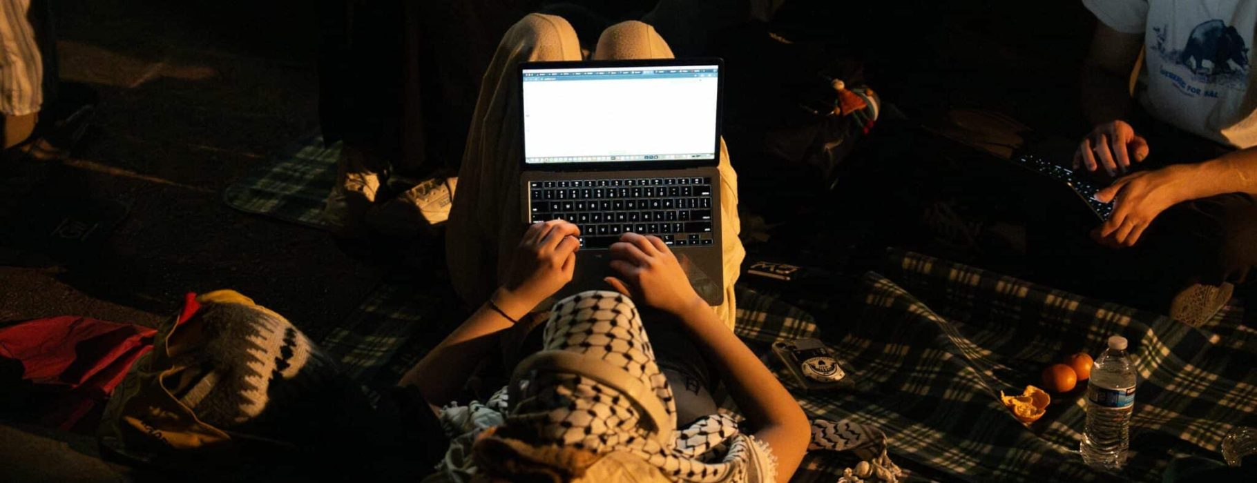 A protester wears a keffiyeh as they work on their laptop in the encampment at night.